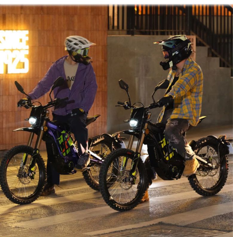 Two riders operating 2026 Sur-Ron Light Bee X 3C street-legal e-motos at night with active front LED headlights and directional indicators in urban terrain.
